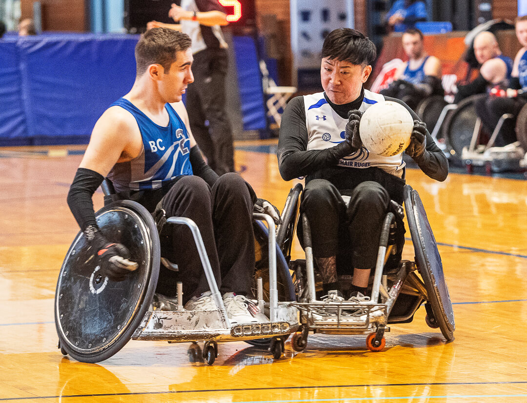 An athlete in a defensive chair protects the ball while being hit by an athlete in an offensive chair.